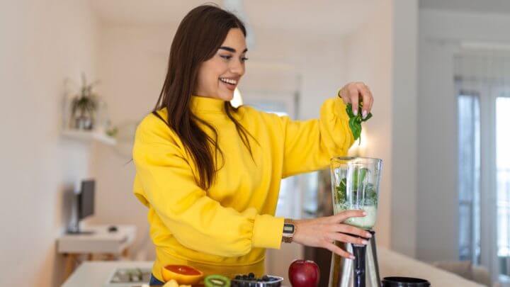 Mulher com blusa amarela na cozinha colocando folhas dentro de liquidificador