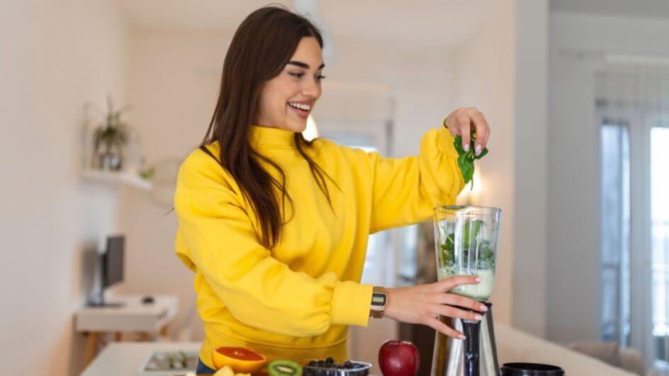 Mulher com blusa amarela na cozinha colocando folhas dentro de liquidificador