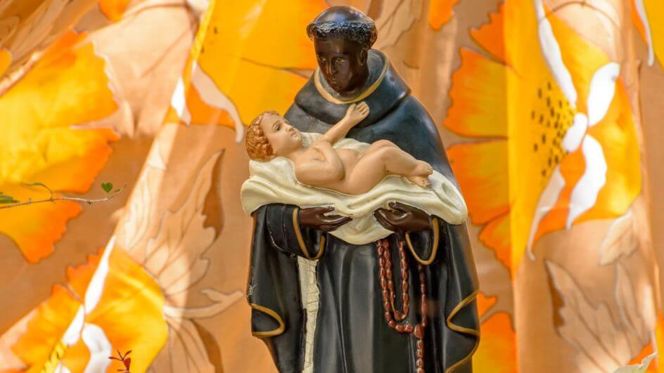 Estátua de São Benedito segurando menino Jesus nos braços e em fundo amarelo