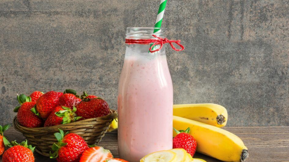 Garrafa de vidro com vitamina de frutas com um canudo dentro. Bananas e morangos inteiros e cortados estão em volta