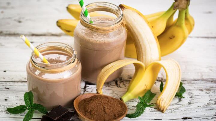 Smoothie de banana com whey sabor chocolate em dois copos separados ao lado de bananas e whey