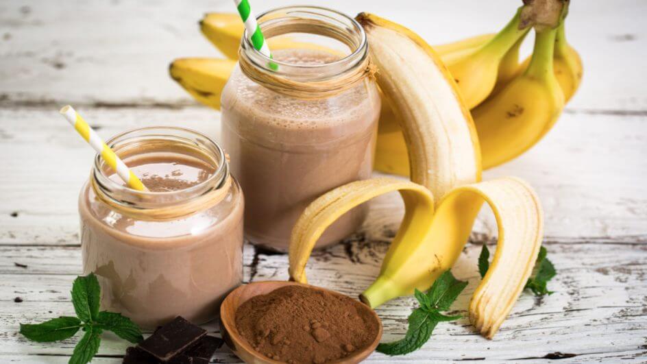 Smoothie de banana com whey sabor chocolate em dois copos separados ao lado de bananas e whey