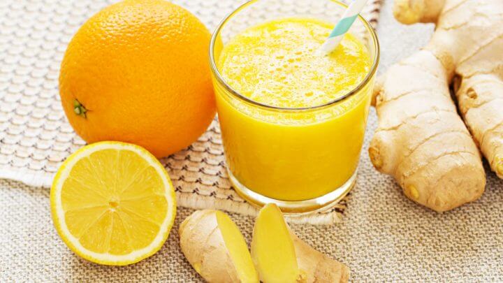 Copo de vidro com suco de laranja com canudo azul e branco. Ao lado do copo tem laranja e gengibre