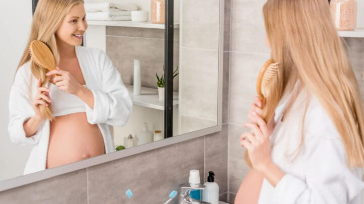 Mulher grávida feliz em roupão escovando o cabelo e olhando no espelho no banheiro