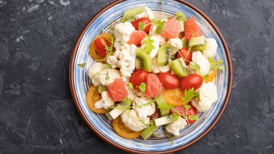 Salada vegetariana de couve-flor, kiwi, tomate, brotos microverdes em fundo de concreto preto; Vista superior, de perto