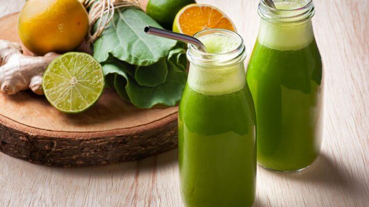 Duas garrafas de suco verde com canudo de metal. Ao lado e ao fundo, couve, laranja e limão