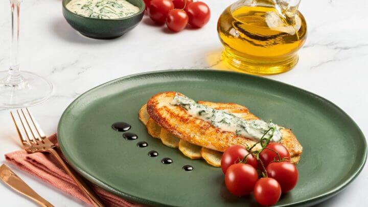 Tilápia com Creme de Espinafre e Redução de Vinho Balsâmico em um prato verde musgo redondo com talheres rosés ao lado, azeite e tomates-cerejas