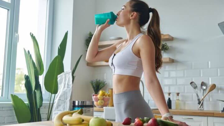 Mulher na cozinha com roupa de academia e bebendo em garrafa verde. Em bancada à frente dela tem diversas frutas