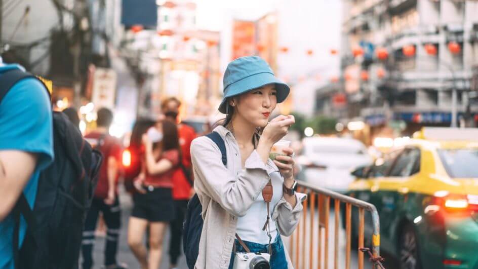 Mulher usando chapéu e com mochila nas costas comendo na rua