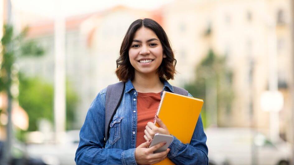 Mulher em pé em uma rua sorrindo e segurando um livro amarelo