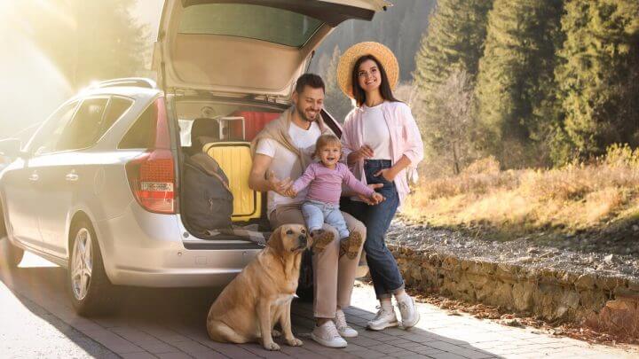 Homem, mulher, bebê e um cachorro em frente ao porta-malas de um carro sentados
