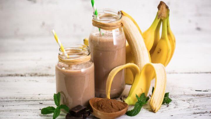 Dois copos com vitamina de banana e chocolate em cima de uma mesa de madeira com bananas e chocolate em pó