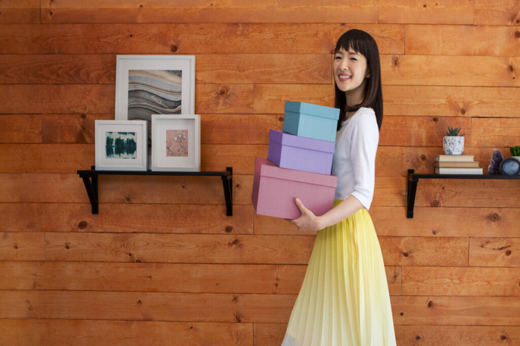 Marie Kondo de lado segurando caixas coloridas
