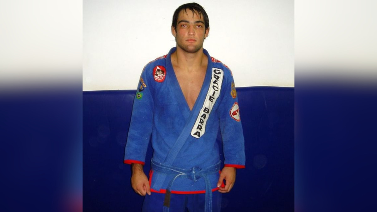 Um homem jovem, vestindo um kimono azul da Gracie Barra, está em pé diante de uma parede. Ele tem cabelo escuro e curto, com uma expressão séria e as mãos relaxadas ao lado do corpo. Há patches de equipe e bandeiras costurados no kimono