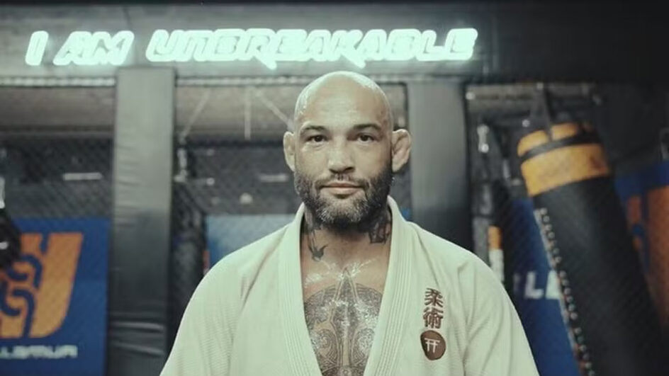 Um homem jovem, vestindo um kimono azul da Gracie Barra, está em pé diante de uma parede. Ele tem cabelo escuro e curto, com uma expressão séria e as mãos relaxadas ao lado do corpo. Há patches de equipe e bandeiras costurados no kimono