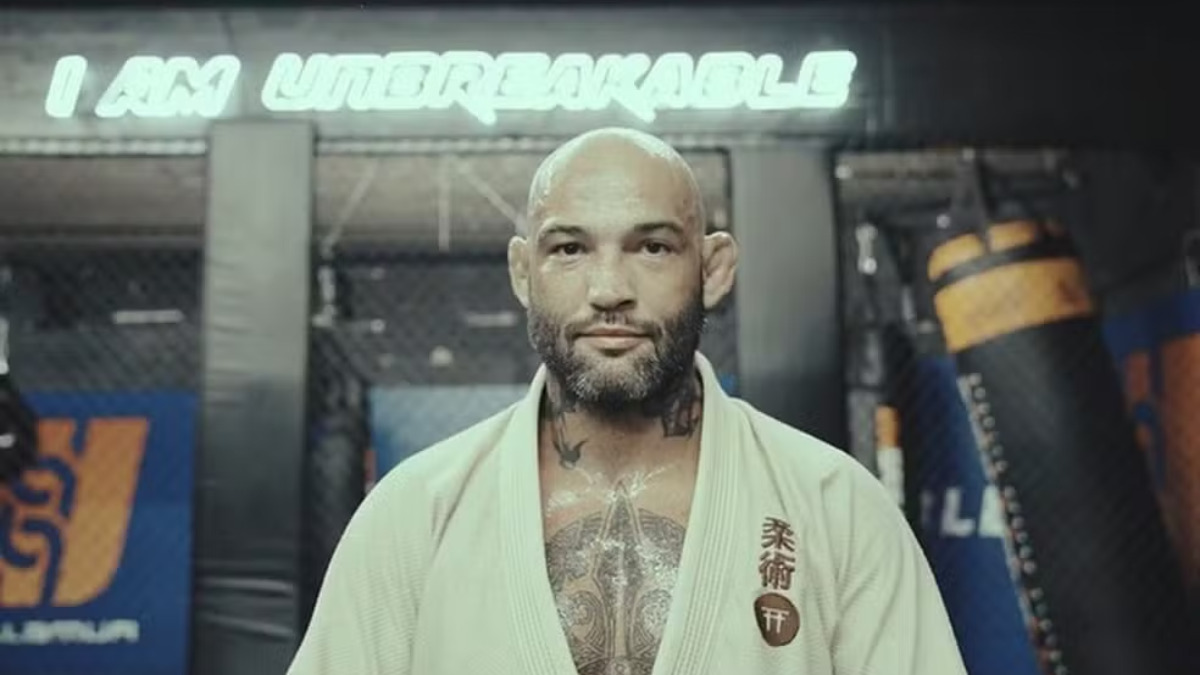 Um homem jovem, vestindo um kimono azul da Gracie Barra, está em pé diante de uma parede. Ele tem cabelo escuro e curto, com uma expressão séria e as mãos relaxadas ao lado do corpo. Há patches de equipe e bandeiras costurados no kimono