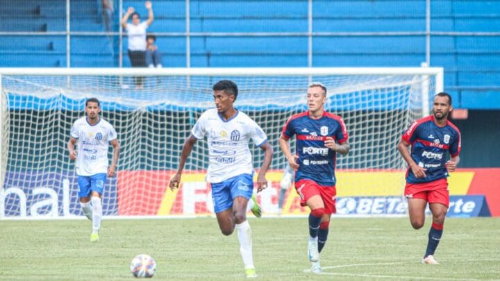Imagem mostra jogador do Caravaggio vestido de camisa branca e acompanhado por jogadores do Marcílio Dias com camisa azul escura e com listra vermelha