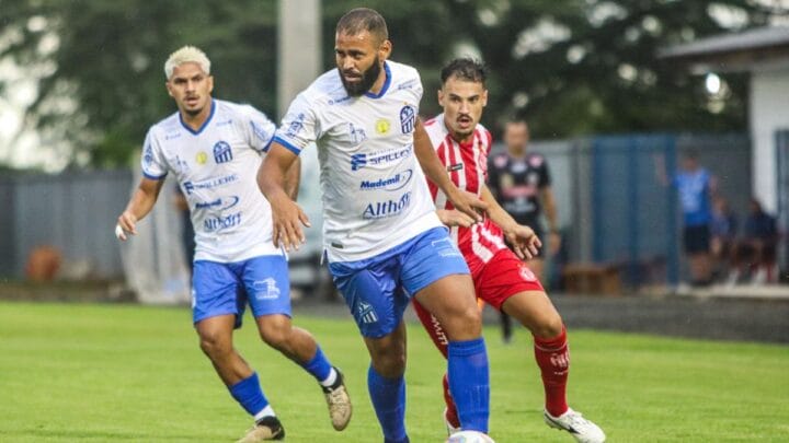 Imagem mostra jogador do Caravaggio com camisa branca e bermuda azul e um jogador do Hercílio Luz no lado direito da foto com o uniforme vermelho e branco
