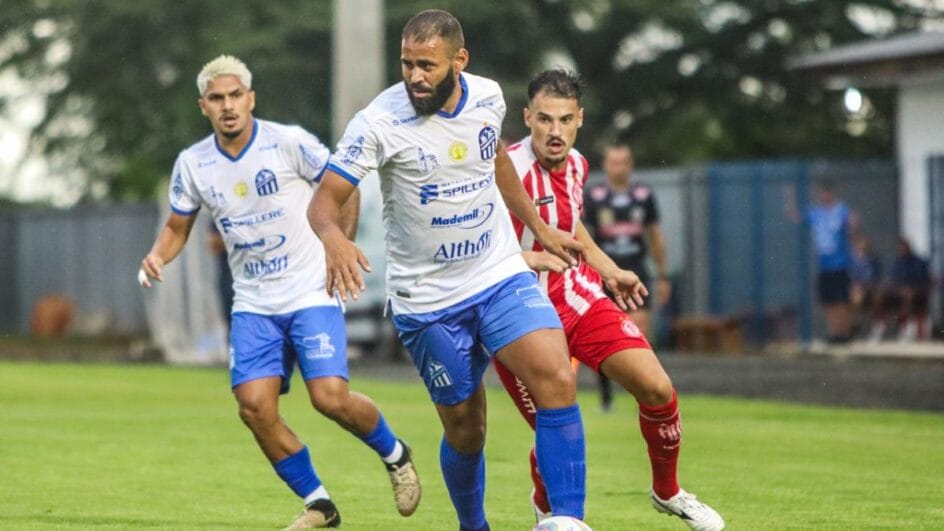 Imagem mostra jogador do Caravaggio com camisa branca e bermuda azul e um jogador do Hercílio Luz no lado direito da foto com o uniforme vermelho e branco