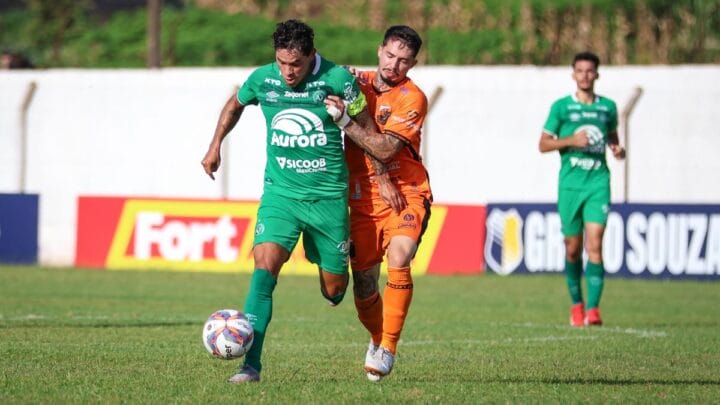 Imagem mostra jogador da Chapecoense de verde e do Santa Catarina de laranja disputando bola em frente do jogador da Chapecoense e o gramado verde
