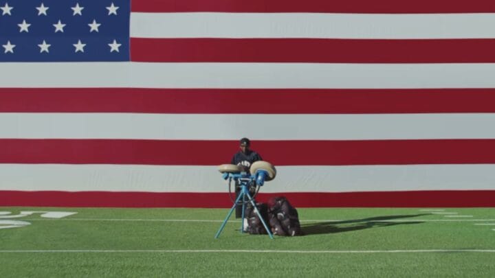 Imagem mostra Kendrick Lamar atrás de uma máquina de lançar bolas no gramado de um campo de futebol americano e ao fundo uma bandeira dos Estados Unidos
