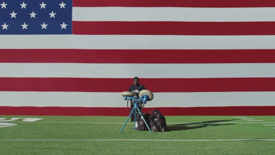 Imagem mostra Kendrick Lamar atrás de uma máquina de lançar bolas no gramado de um campo de futebol americano e ao fundo uma bandeira dos Estados Unidos