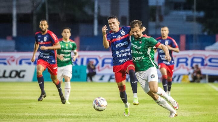 Imagem mostra jogador do Concórdia vestido de camisa verde e do Marcílio Dias com camisa azul disputando bola