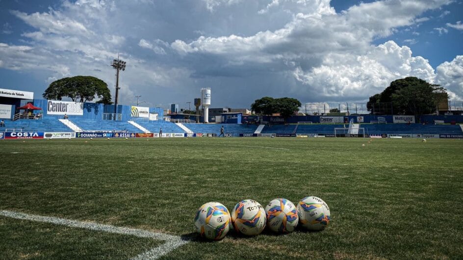 Imagem mostra o gramado do Estádio Anibal Batista e quatro bolas alinhadas. O céu aparece com muitas nuvens, mas está de dia