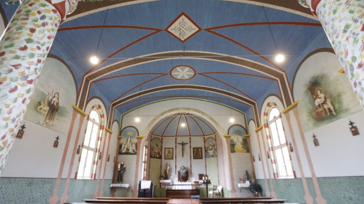 Interior da Capela Nossa Senhora do Perpétuo Socorro, em Indaial (SC), com teto azul decorado, pinturas coloridas, altar ao fundo e arquitetura única tombada pelo Iphan.
