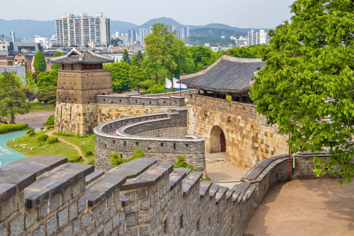 Vista da Fortaleza Hwaseong, com um muro de pedras cinza, muralha bege e uma cidade ao fundo
