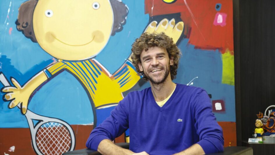 Imagem mostra Guga Kuerten sentado com uma camisa azul e ao fundo um desenho
