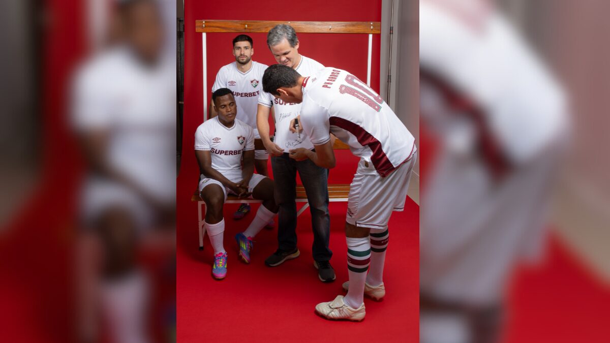 Martinelli, Jhon Arias e PH Ganso assinando o novo uniforme do Fluminense de torcedor presenteado - (Foto: Umbro e Lucas Merçon, FFC)