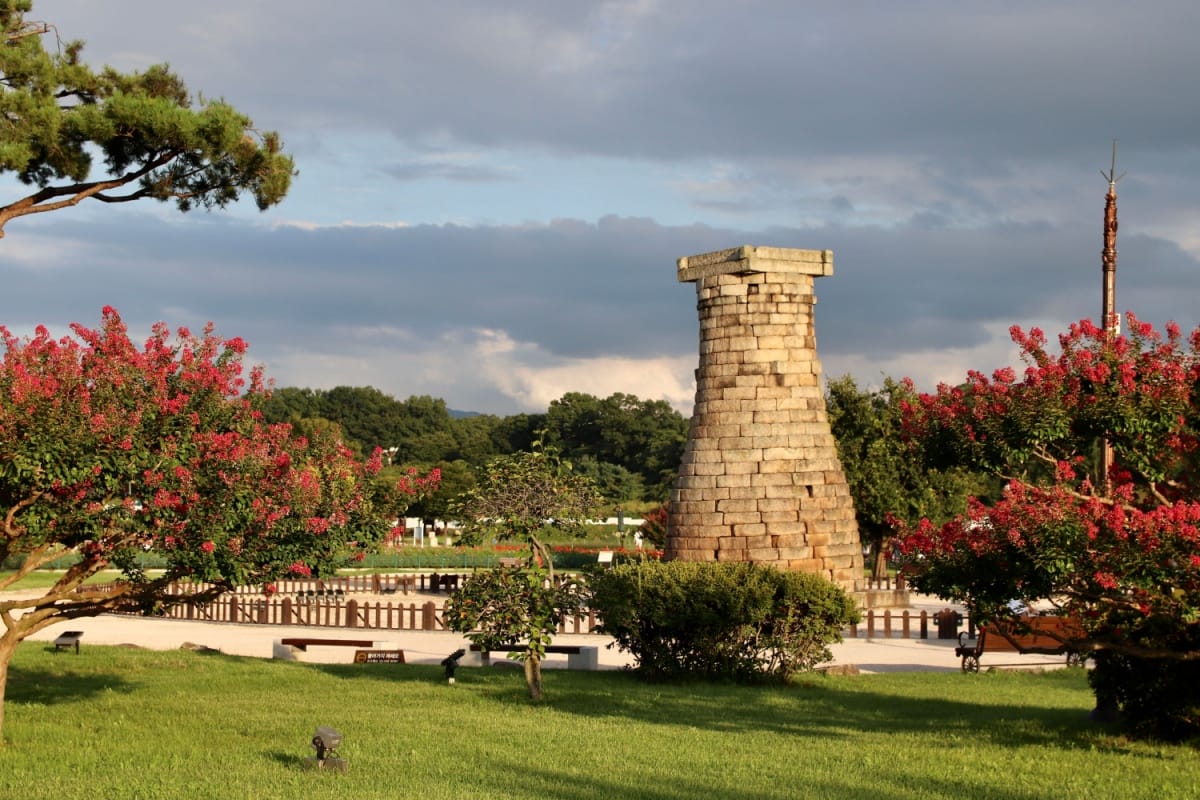 Vista do Observatório Cheomseongdae, com uma muralha e um jardim com gramado verde e árvores