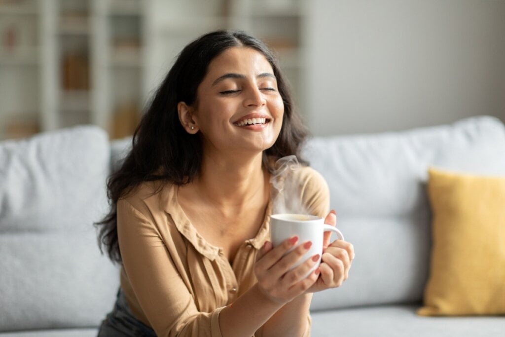 Mulher sorrindo com os olhos fechados, sentada em um sofá, e segurando uma xÃcara de café