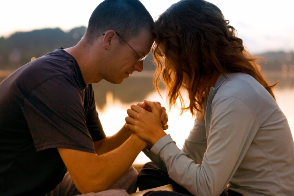 Homem de cabelo curto usando oculos e camiseta preta de mÃ£os dadas e com a testa encostada em mulher com o cabelo curto, usando uma camisa de manga longa cinza.