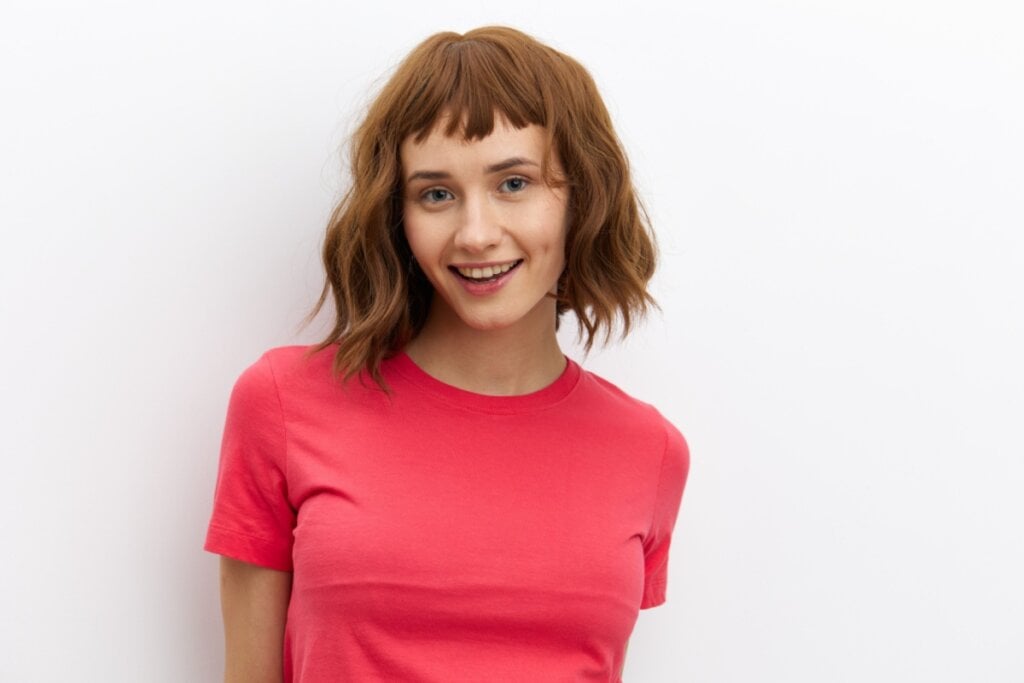 Mulher com cabelo ruivo curto com franja curta, sorrindo e usando uma blusa rosa