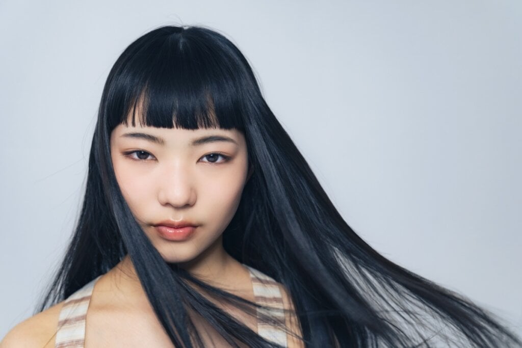 Mulher com cabelo longo preto com franja reta posando para foto em um fundo cinza