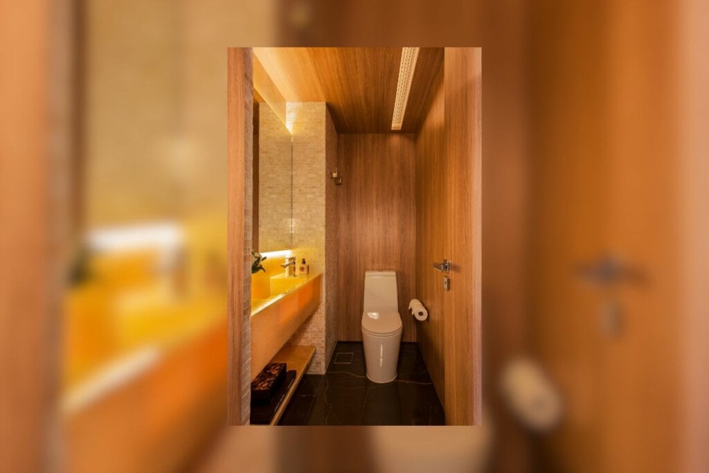 Banheiro com revestimento de madeira, luz amarela, espelho na lateral e uma privada branca ao fundo
