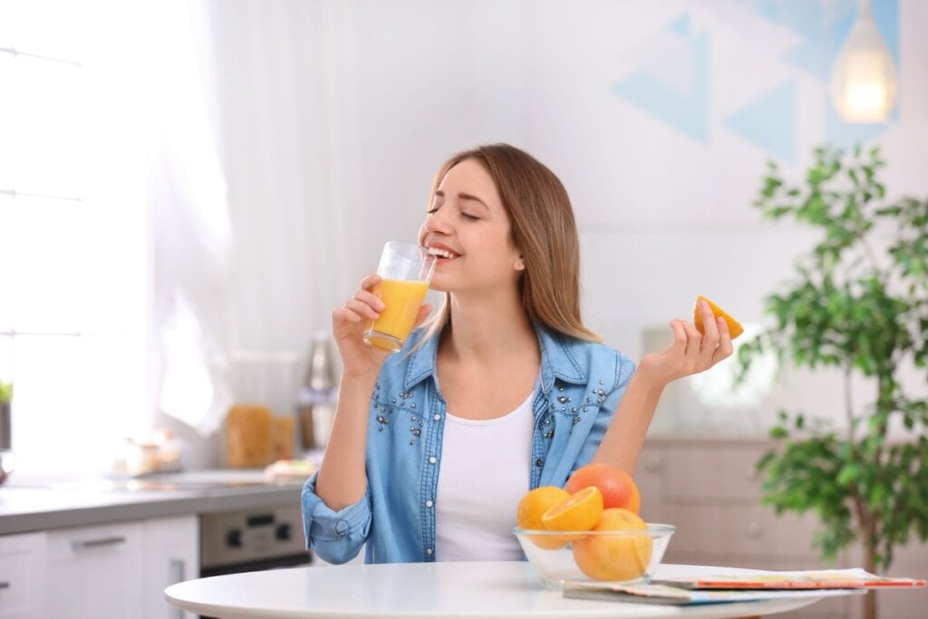 Mulher sentada em bancada tomando suco de laranja, ela estÃ¡ vestindo uma camiseta branca e uma camisa azul.