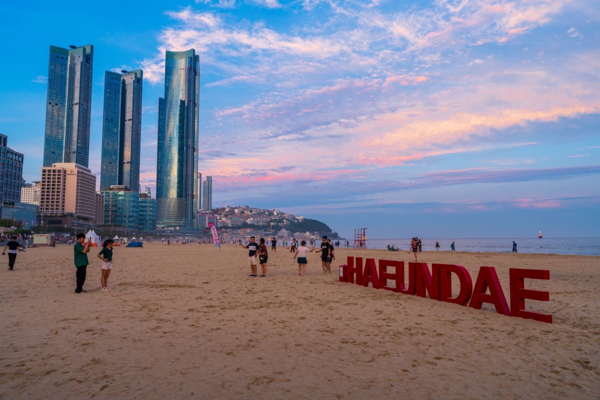 Vista da praia de Haeundae no entardecer, com céu azul, rosa e laranja, e pessoas andando na faixa de areia
