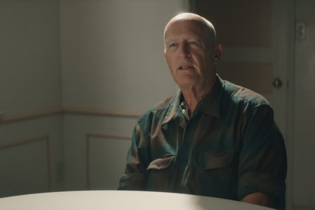 Homem careca usando uma roupa marrom e verde sentado em frente a uma mesa