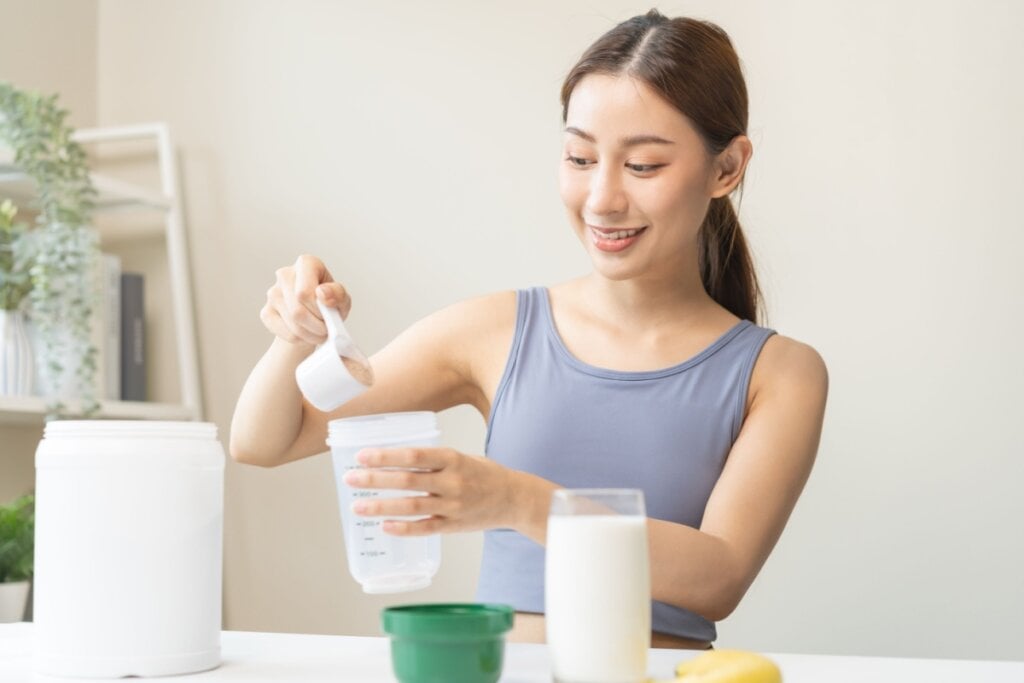 Jovem asiática com o cabelo preso em rabo de cavalo, usando uma regata roxa preparando shake de proteÃna