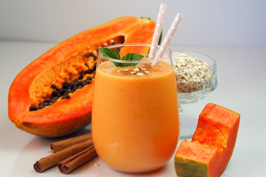 Vitamina de mamÃ£o com aveia e canela servido em copo de vidro, decorado com hortelÃ£ e canudos, ao lado de mamÃ£o, pau de canela e aveia em flocos