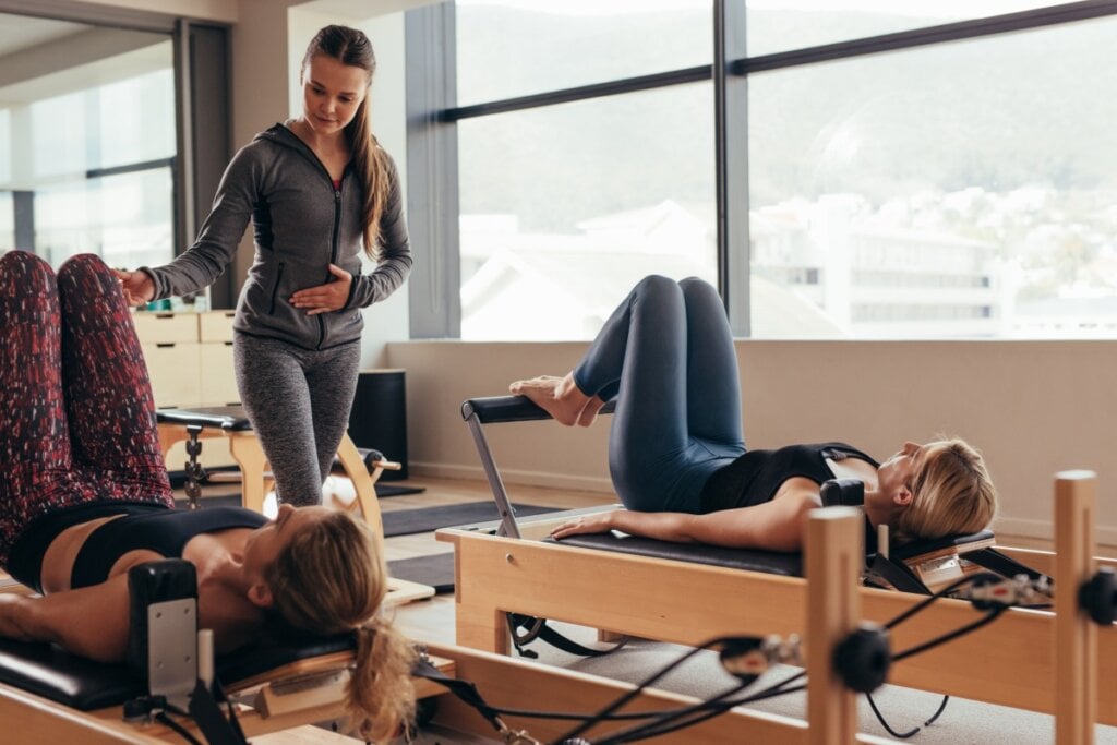 Duas mulheres com roupas de academia deitadas em aparelho realizando exercÃcio de pilates acompanhadas de professora de pilates usando roupas de academia na cor cinza