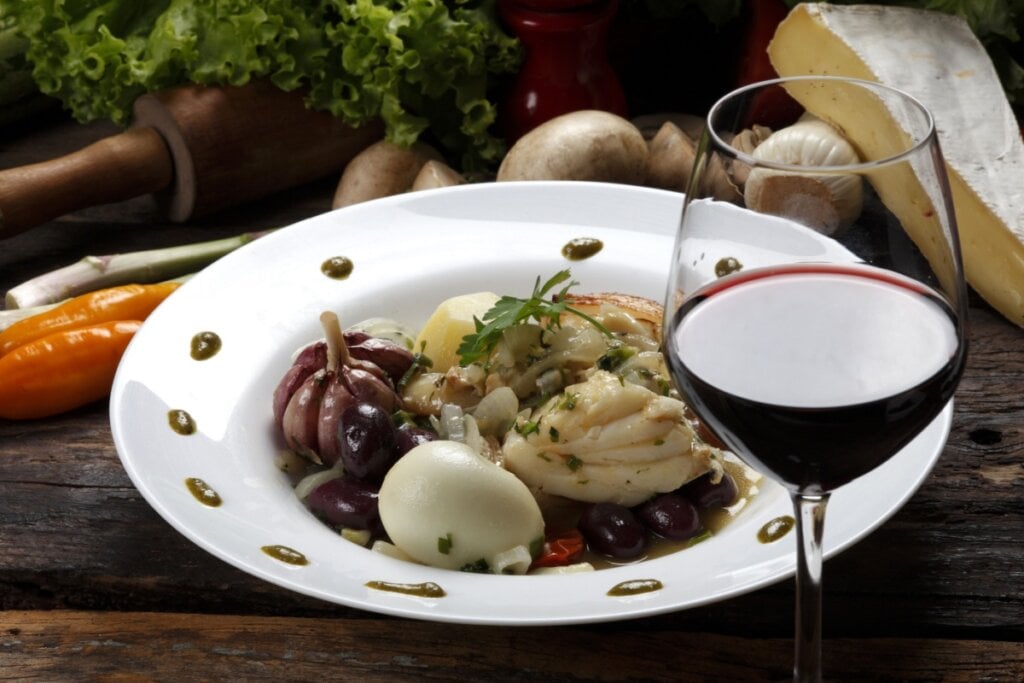 Prato com bacalhau acompanhado de legumes e taÃ§a de vinho tinto em uma mesa rÃºstica com ingredientes ao fundo