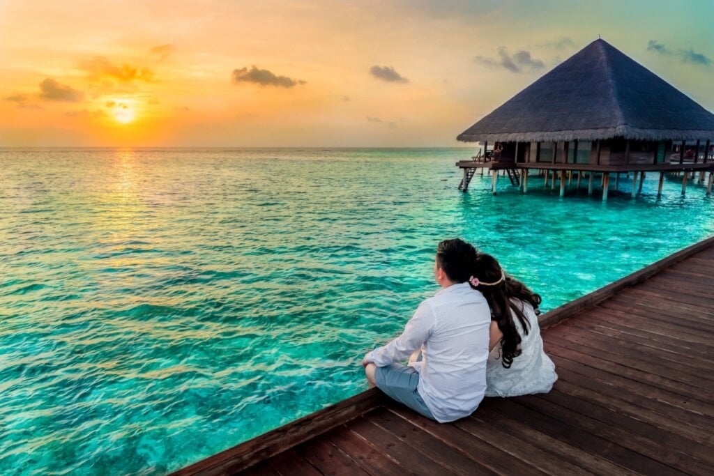Casal sentado em um deck de madeira assistindo ao p&Atilde;&acute;r do sol diante do mar azul-turquesa nas Maldivas ao lado de bangal&Atilde;&acute;