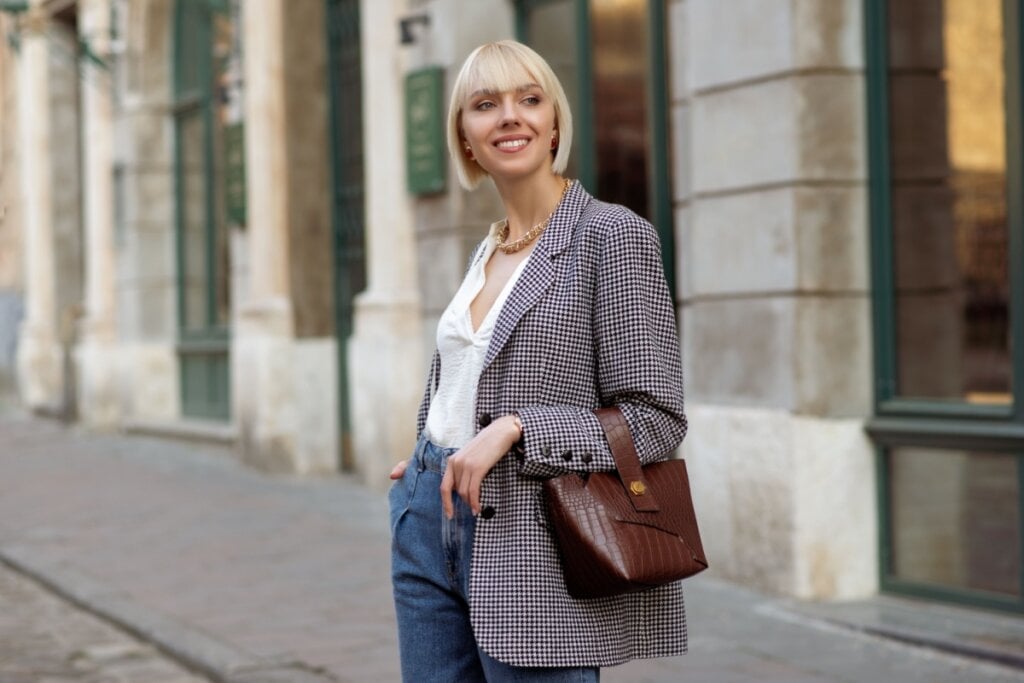 Mulher loira com blazer xadrez, blusa branca e calça jeans sorrindo em rua