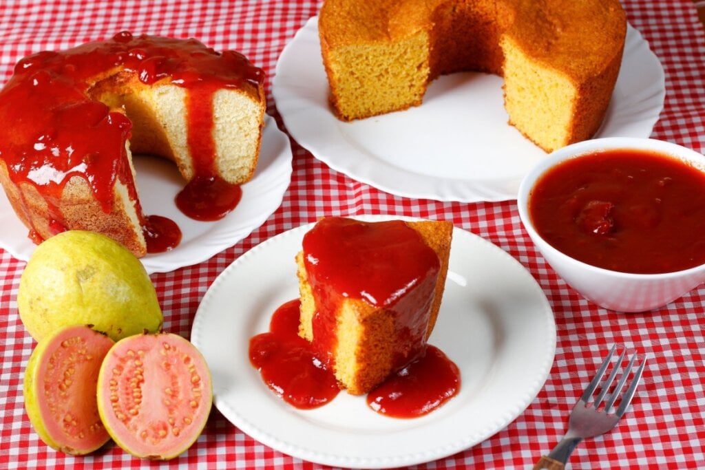 Fatia de bolo de fubá coberta com calda de goiabada ao lado da fruta cortada, bolos cortados e tigela com cobertura sobre toalha xadrez vermelha