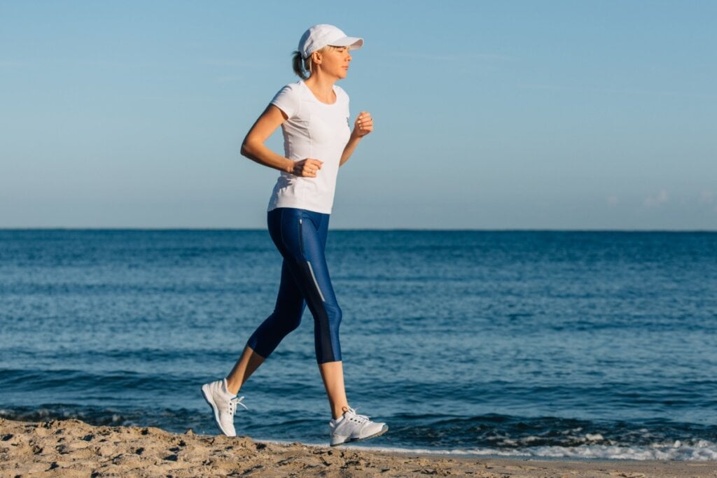 Mulher correndo na areia, usando um bonÃ© e camiseta branca, e uma calÃ§a azul e tÃªnis branco