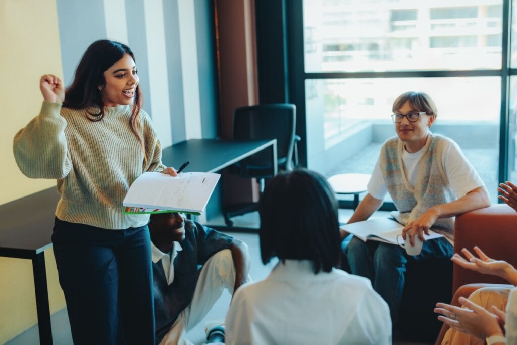 Mulher em pÃ© segurando um caderno e falando para pessoas sentadas